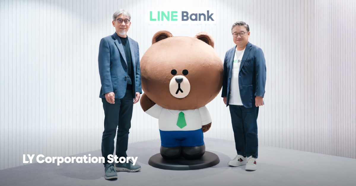 Morris and Fan are standing on either side of a large bear character statue in front of a white vertically striped backdrop featuring the 