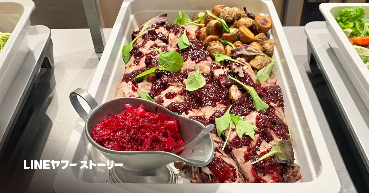 中央にはデンマーク料理のフレスケスタイが大皿に盛られており、スライスされた豚肉に濃い赤色のベリーソースがたっぷりとかかり、上に緑の葉野菜が散らされている。右側には丸ごとの小さなジャガイモが添えられ、手前には赤いキャベツの付け合わせが銀色のソースボートに入って置かれている。