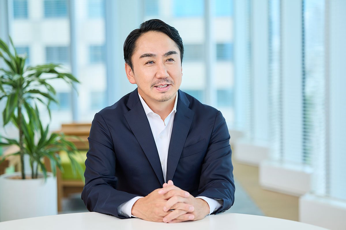 スーツ姿のCEO出澤が、テーブルに両手を置いて笑顔で話している写真