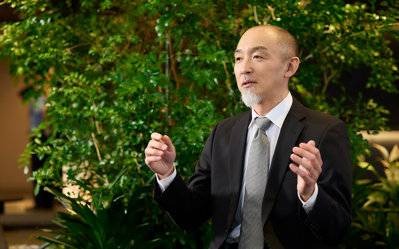 鈴木が両手を肩の高さに上げて間隔を示すように広げながら説明している。視線は前方に向けられ、落ち着いた表情で話している様子