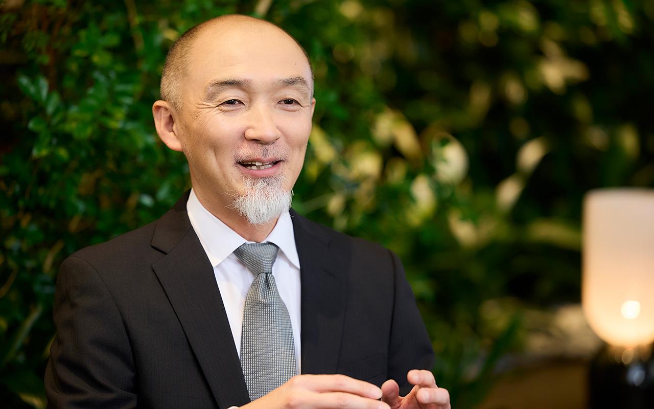 鈴木が両手を軽く前に出しながら会話している。口元に笑みを浮かべ、リラックスした表情で相手に視線を向けている様子