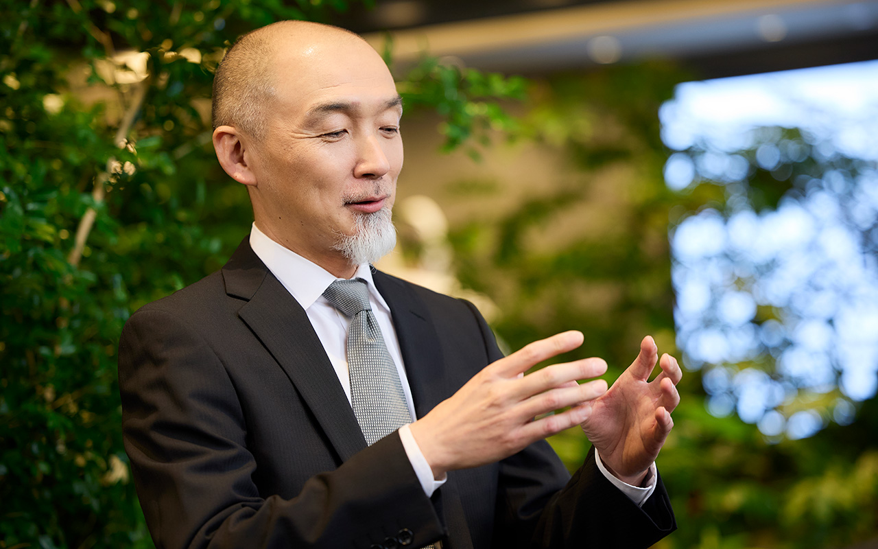 鈴木が両手を前に出して大きさや形を示すように動かしながら説明している。視線は手元付近に向けられ、穏やかな表情で会話している様子