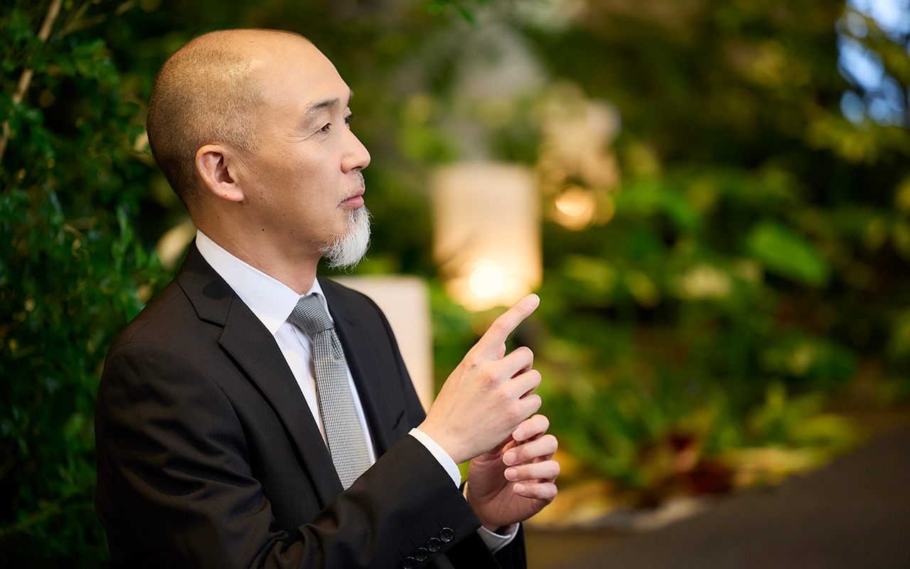 鈴木が横を向いて右手の人差し指を上げてポイントを示すようにしながら説明している。視線は前方に向けられ、落ち着いた表情で話している様子