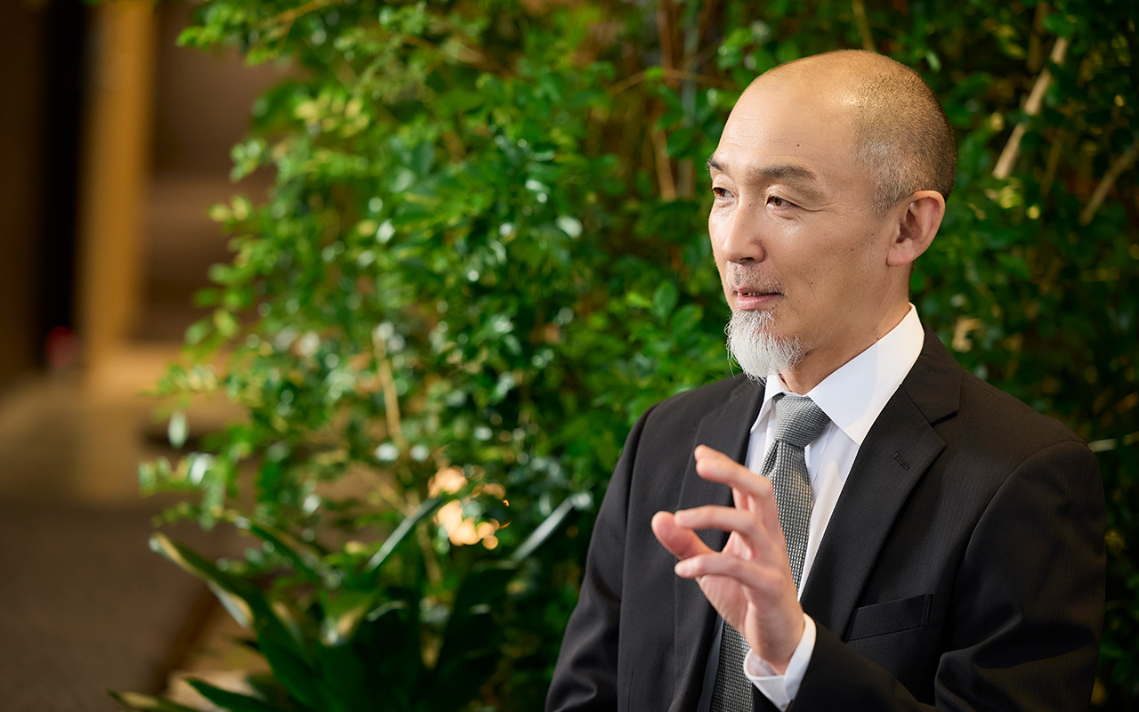 鈴木が植物が多く配置された室内で横向きに座り、右手の指を軽くつまむように上げながら説明している。視線は前方に向けられ、落ち着いた表情で会話している様子