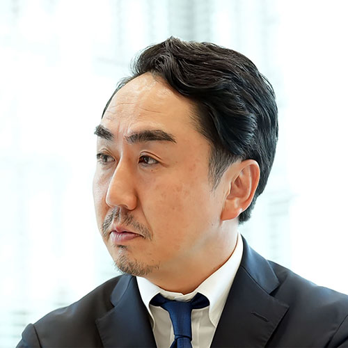 スーツを着た出澤が横を向いて考え込んでいます。背景の窓から自然光が差し込み、落ち着いた雰囲気です。