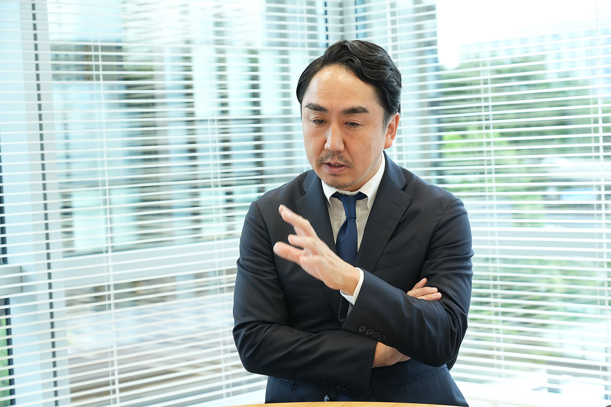 スーツ姿の出澤が腕を組み、手振りで要点を示しながら説明しています。背後に窓のブラインドと外の景色が写る、会議室での発言シーンです。