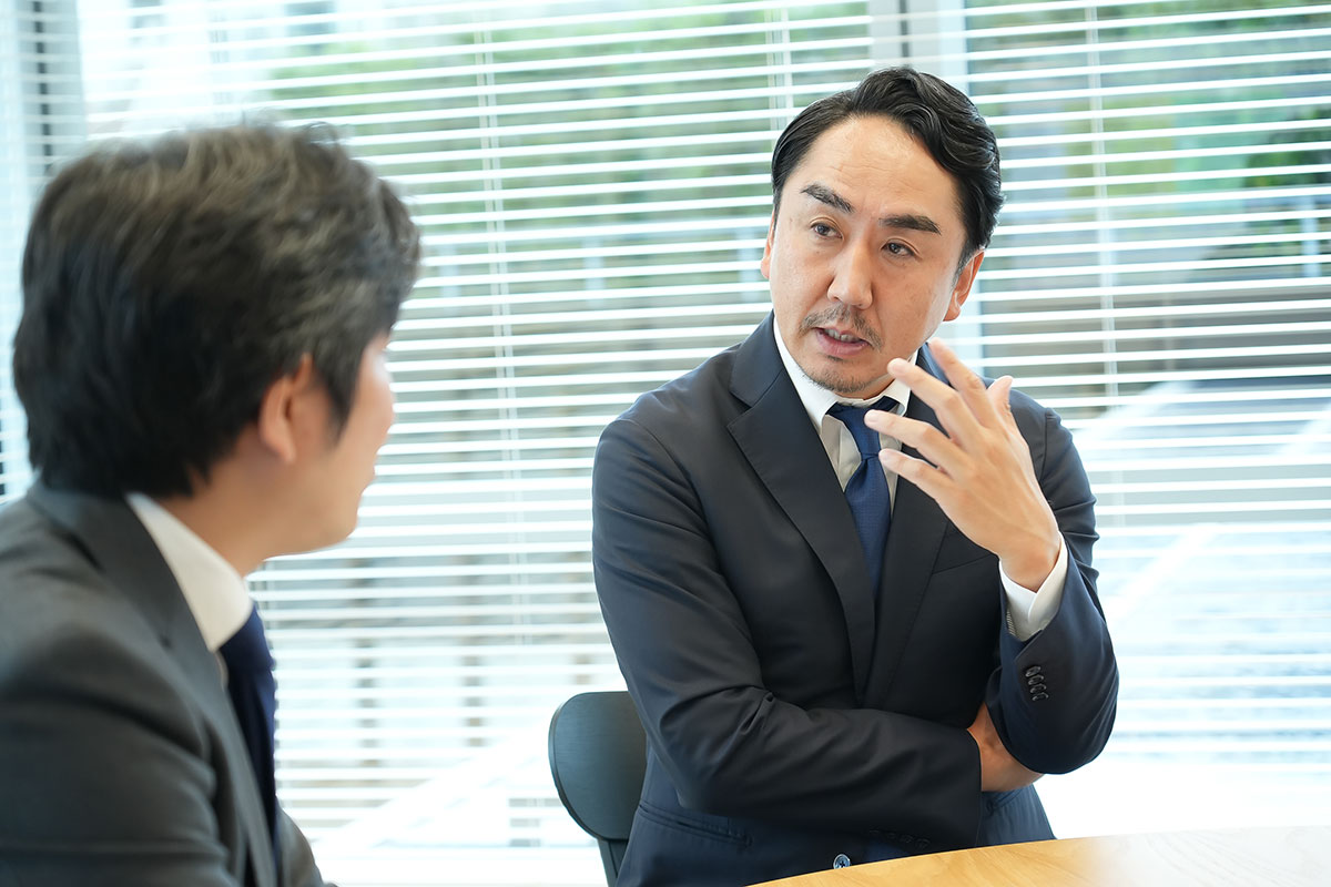 会議室の丸テーブルで、スーツ姿の出澤が身振りを交えて説明し、向かいの男性が聞いています。ブラインド越しに屋外が見える打合せの場面です