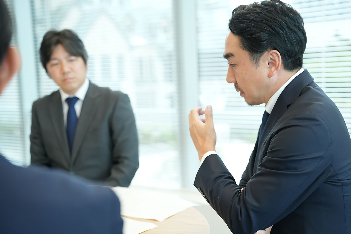 会議の様子。手前の人物がぼかされ、奥のスーツ姿の出澤がジェスチャーをしながら話しています。丸テーブルと資料が写るディスカッションの場面です