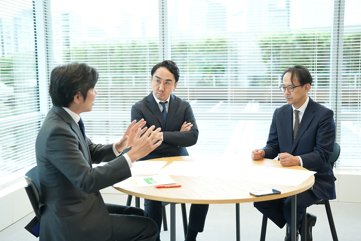 会議室の丸テーブルで、スーツ姿の出澤、川邊、妹尾が議論をしています。資料が机上に置かれ、窓のブラインド越しに外の景色が見えます