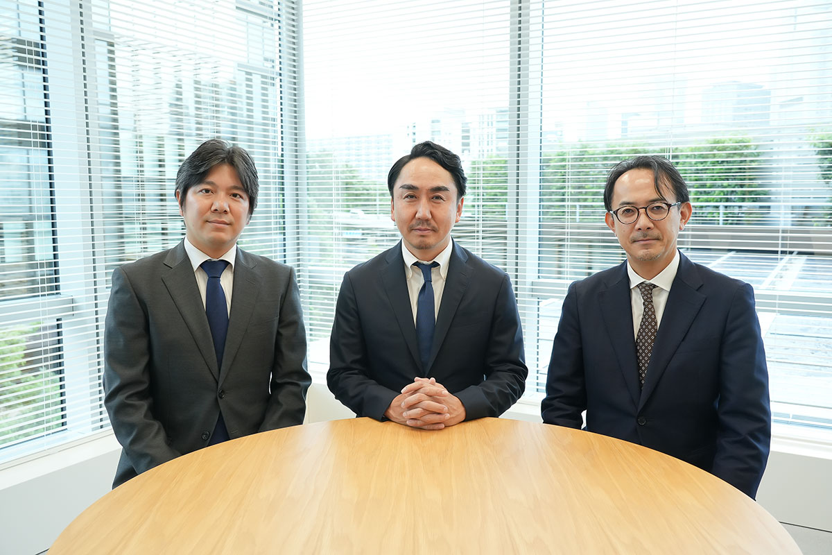 窓のブラインドを背景に、スーツ姿の出澤、川邊、妹尾が丸テーブルの前に並んで座っています。会議室での集合カットで、正面を向いて撮影されています。