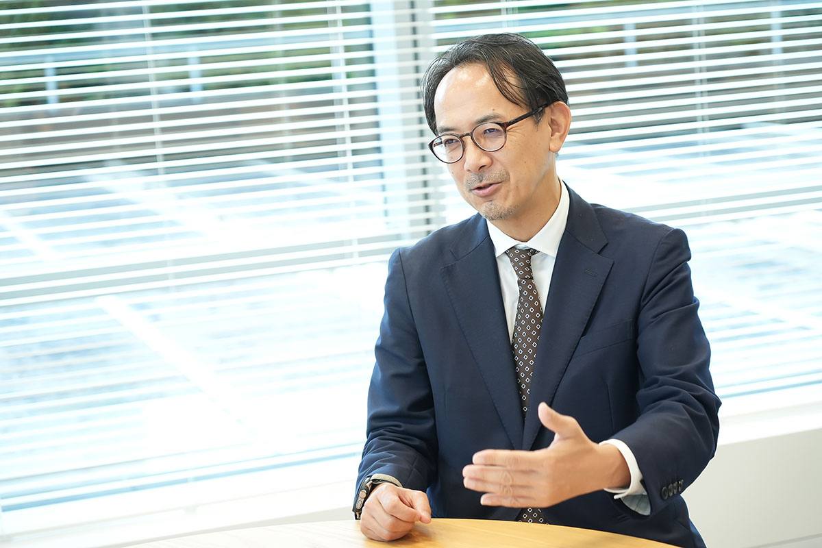 眼鏡をかけたスーツ姿の川邊が、手を前に出して発言しています。丸テーブルに資料が置かれ、ブラインド越しに背景が見える会議中の場面です。