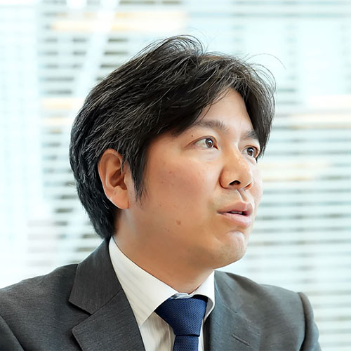 スーツ姿の妹尾が横を向いて話しています。背景にはブラインドがあり、真剣な表情で何かを説明している様子です
