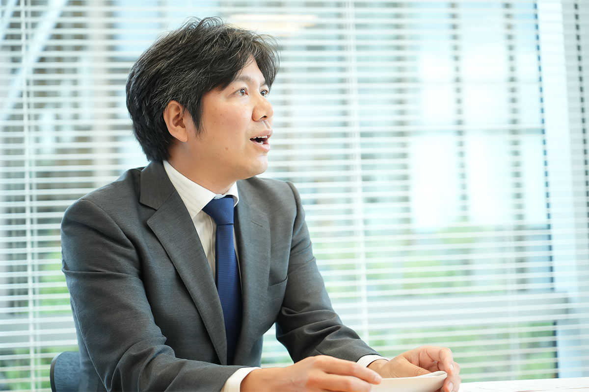 ブラインドの前で、スーツ姿の妹尾が斜め方向に視線を向けて発言しています。テーブルの資料に手を添えた姿が写る、会議中の一場面です
