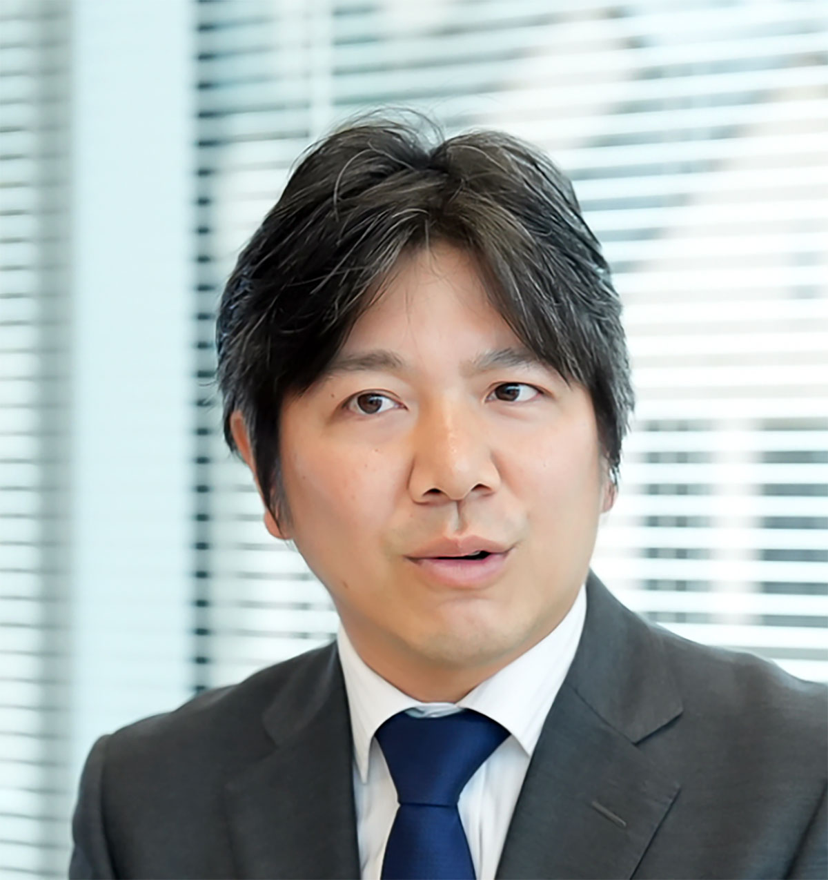 スーツ姿の妹尾が話しています。背景にはブラインドがあり、真剣な表情で何かを説明している様子です
