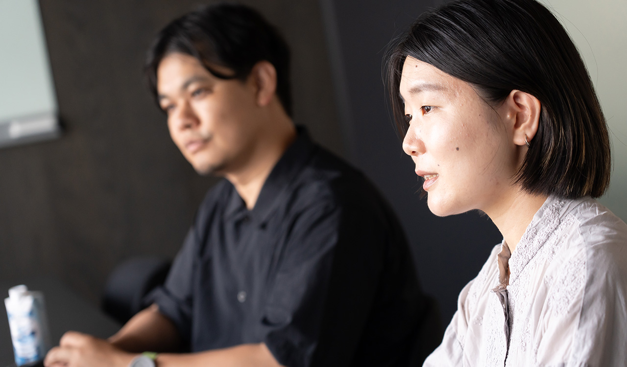 金子さんのバストショット。淡いグレーの長袖ブラウスを着用し、あごのラインでそろえた黒髪のボブヘアで、横向きに座って話している様子