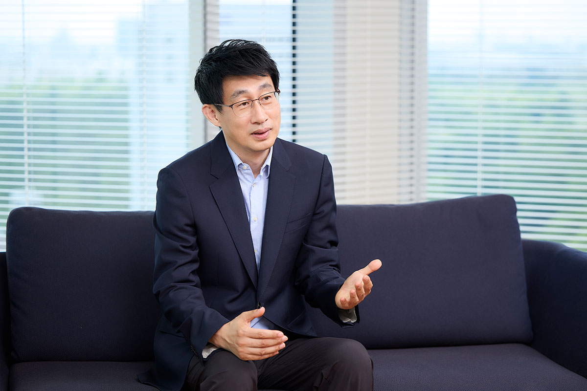 Shin, a man in a suit, is sitting on a sofa, using his hands to explain something. Natural light is streaming in through the window.