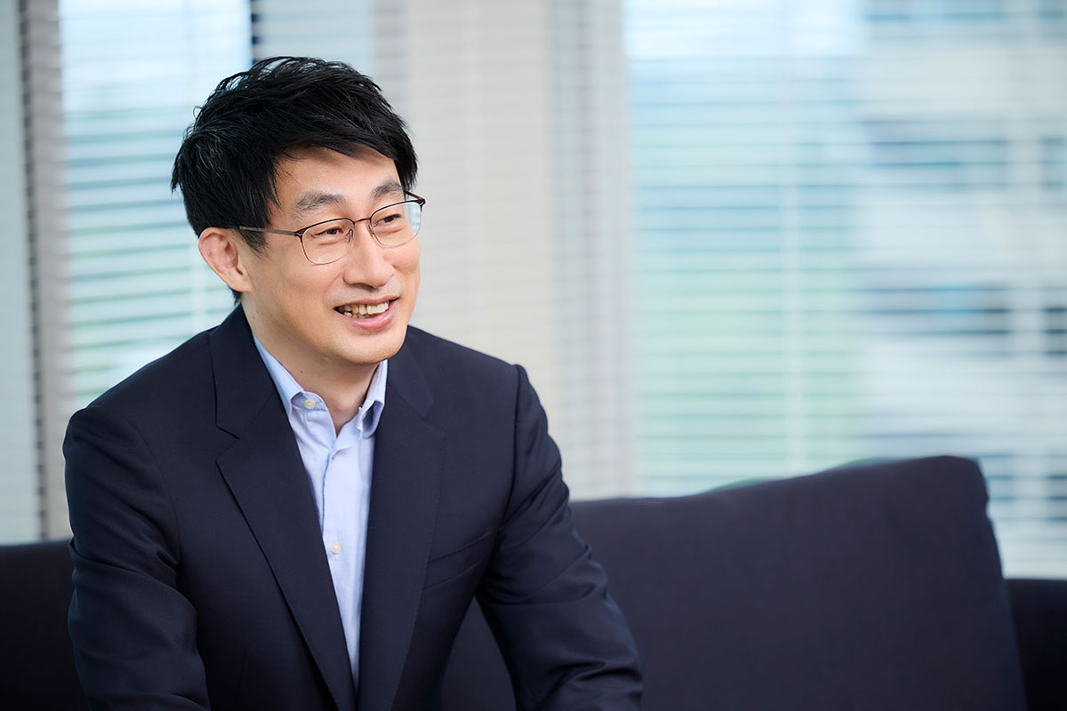 Shin, dressed in a suit, is speaking with a gentle smile. Light pours in through the window, creating a relaxed atmosphere.