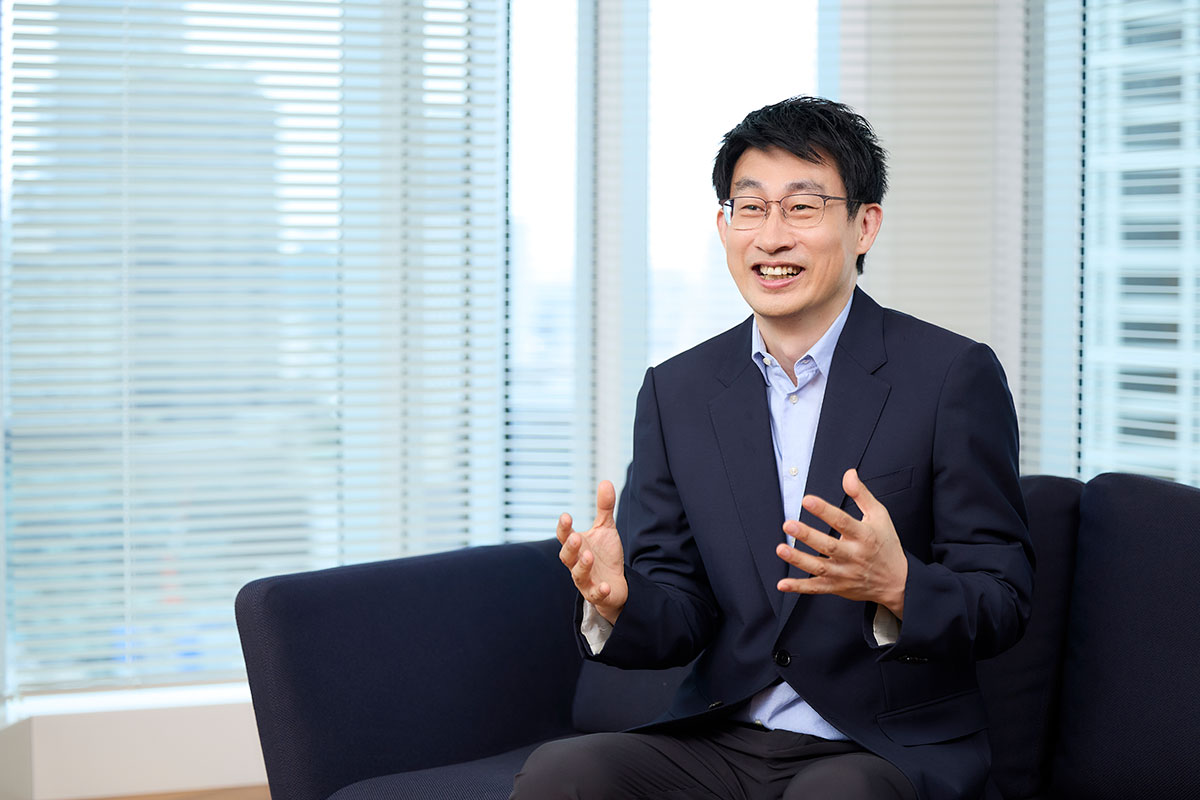 Shin, dressed in a suit, is sitting on a sofa, speaking with his hands open and a cheerful expression. Blinds can be seen in the background.