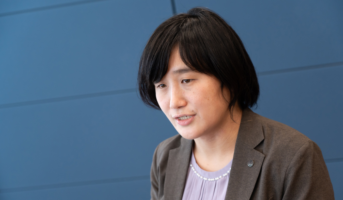 警察庁の青田さんのバストショット。青田さんは黒髪のショートヘアで、グレーのジャケットに淡い紫色のブラウスを着ている。やや下を向きながら口を開いており、話している様子。