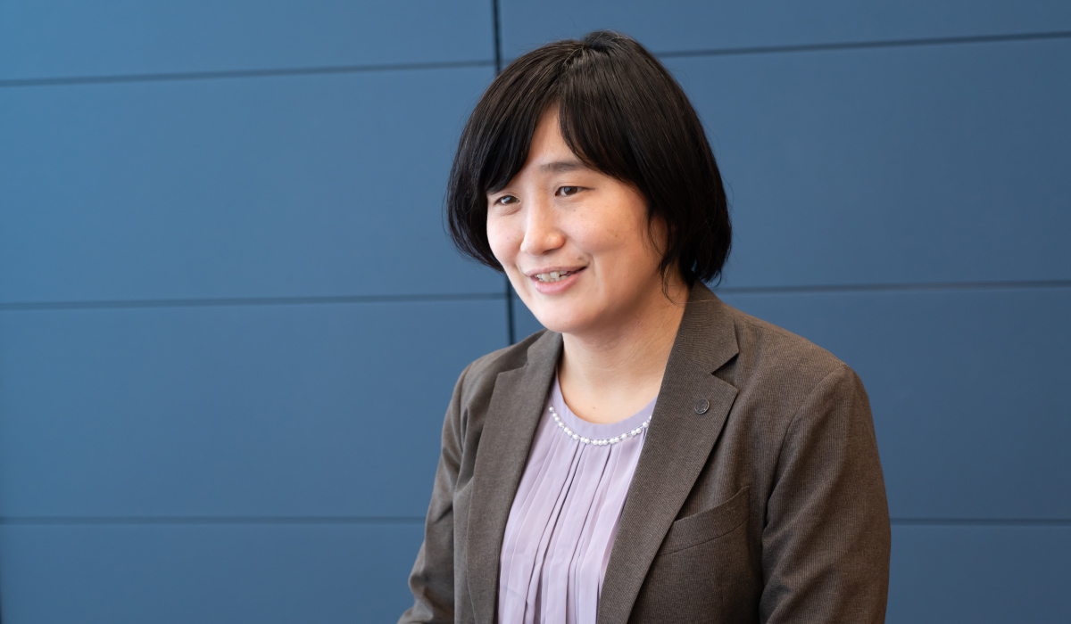 警察庁の青田さんのバストショット。青田さんは黒髪のショートヘアで、グレーのジャケットに淡い紫色のブラウスを着ている。やや右方向を向いて笑顔を見せている。