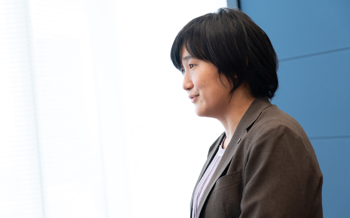 警察庁の青田さんの横顔ショット。軽く笑みを浮かべながら話している様子。背景は右側が青い壁、左側は白く明るい窓辺で、光が強く差し込んでいる。