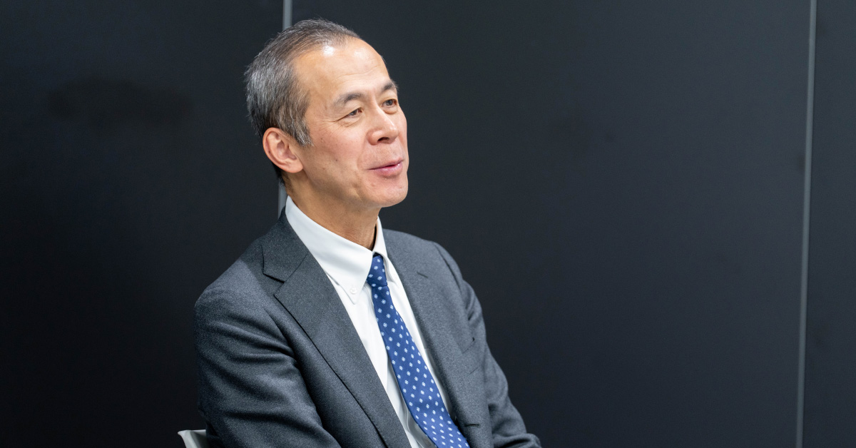 櫻澤さんの上半身ショット。右方向を向いて微笑みながら話している様子。背景は黒っぽい壁で、落ち着いた印象を与えている。