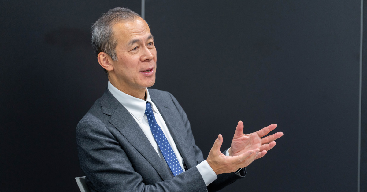 櫻澤さんの上半身ショット。両手を広げて会話の中で説明をしている様子。表情は柔らかく、背景は黒い壁で落ち着いた雰囲気。