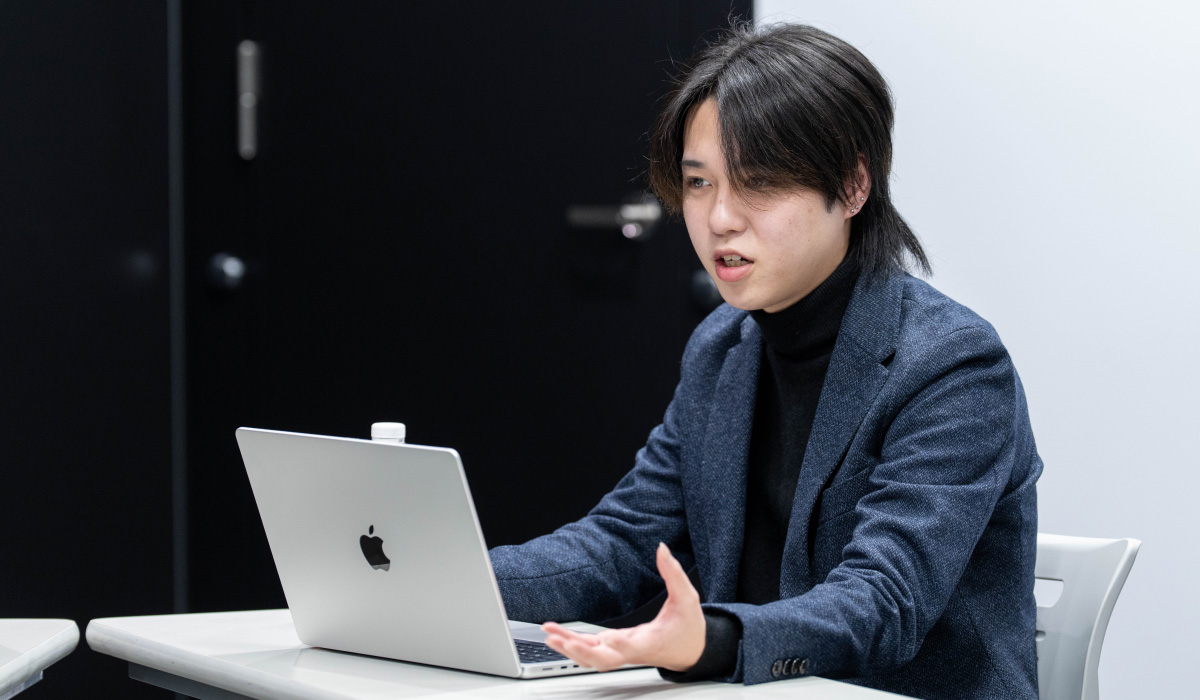 首浦の上半身ショット。白いテーブルにMacBookを置いて操作しながら、左手を前に出しつつ話している様子。背景は黒と白の壁で、真剣に話している。