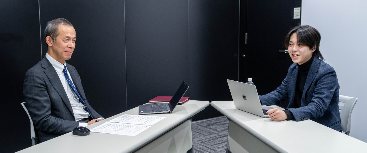 櫻澤さんと首浦が向かい合って座っている会議室の様子。2人の間にはL字型に配置された白いデスクがある。