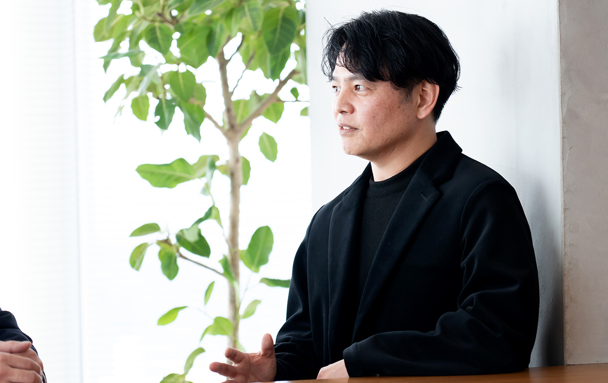 LINEヤフーの西田さんのバストショットである。黒いジャケットに黒のインナーを合わせ、黒髪を自然に横分けにしている。椅子に座り、体をやや横に向けながら、両手を前に出して軽く動かしつつ話している様子である。