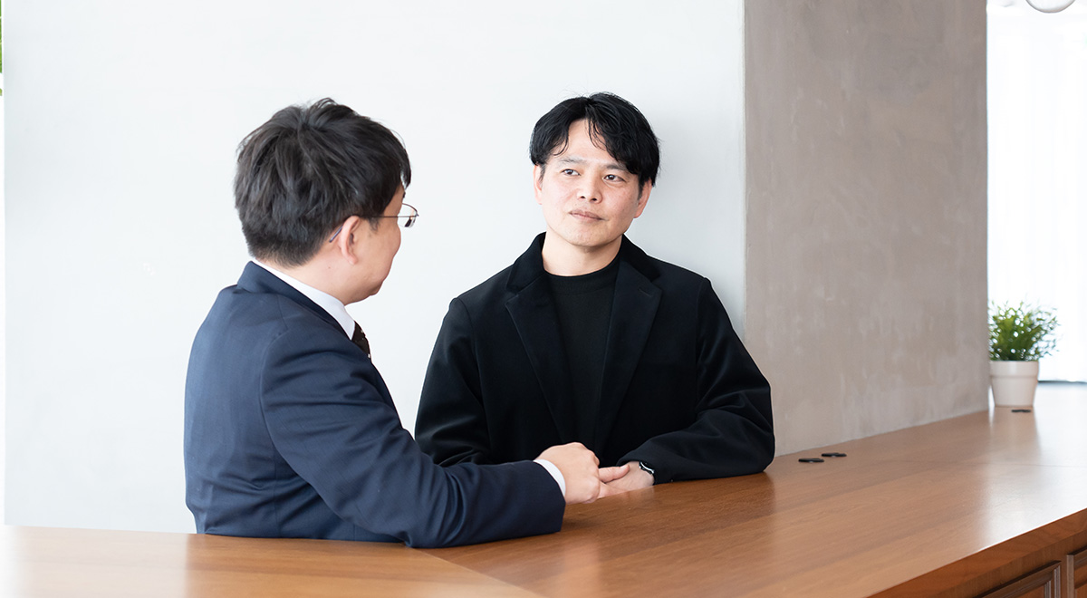 静岡大学の塩田先生とLINEヤフーの西田さんが向かい合って着席している写真である。西田さんは黒いジャケットに黒のインナーを合わせ、自然な黒髪を横分けにしている。両手を軽く組み、相手の話を落ち着いた表情で聞いている。