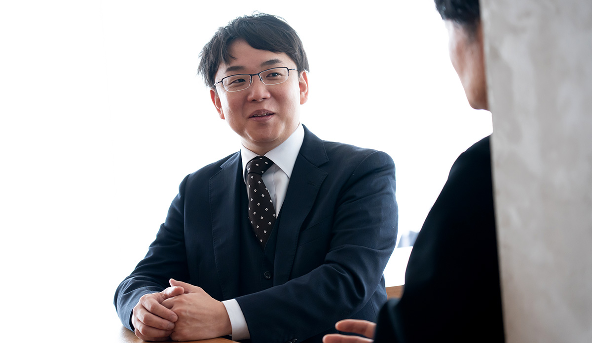 静岡大学の塩田先生のバストショットである。黒いスーツに白いシャツ、黒地に小さなドット柄のネクタイを着用し、短く整えた黒髪で眼鏡をかけている。両手をテーブルの上で組みながら、やや身を乗り出して対面の相手に向かって話している様子。表情は穏やかで、視線は右側にいる相手に向けられている。