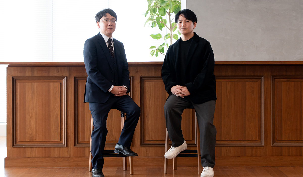 静岡大学の塩田先生とLINEヤフーの西田さんがハイチェアに腰掛けている全身写真。二人は木目調のカウンターの前に並んで座り、背後には観葉植物と明るい窓、木製パネルの壁面が広がる室内空間となっている。