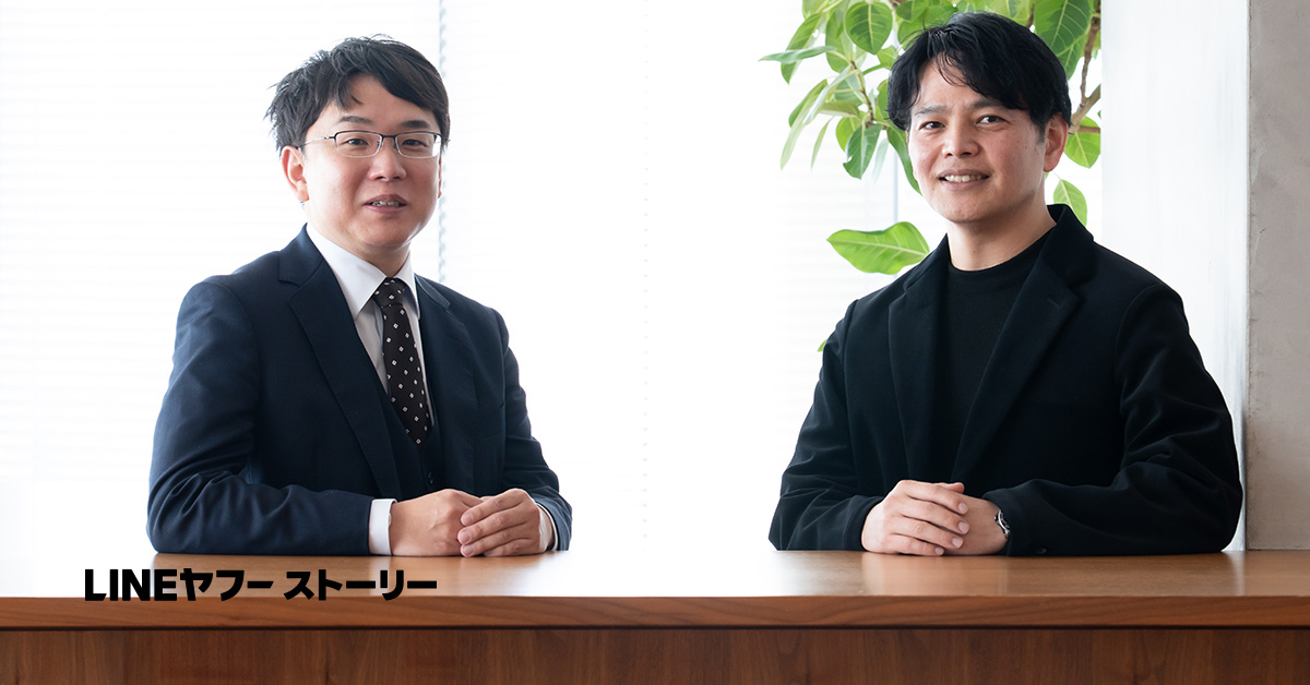 静岡大学の塩田先生とLINEヤフーの西田さんのバストショット。塩田先生は黒いスーツに白いシャツ、黒地に小さなドット柄のネクタイを着用し、短く整えた黒髪で眼鏡をかけている。西田さんは黒いジャケットに黒のインナーを合わせ、自然な黒髪を横分けにしている。両手をテーブルの上に重ね、柔らかく微笑みながら正面を向いている。