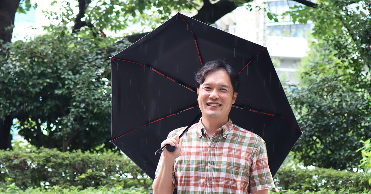 黒い傘を差し、微笑む河津さんが緑豊かな場所でポーズをとっています。雨の中でも明るい雰囲気です。