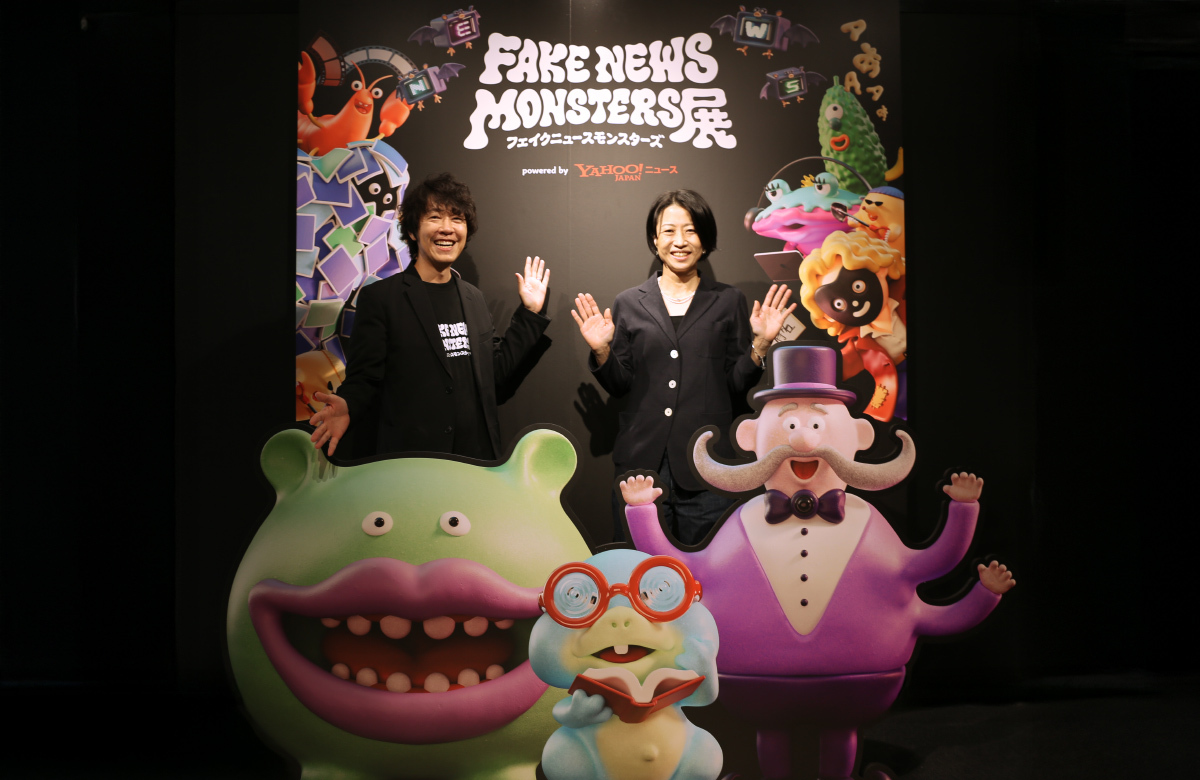 「FAKE NEWS MONSTERS展」の看板前で、男女2人が笑顔で手を上げて立っている。前にはカラフルなモンスターの立体パネルが並んでいる