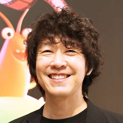 男性のバストショット。黒いジャケットと黒いTシャツを着ており、笑顔でカメラを見つめている。背景にはカラフルなモンスターのイラストがぼんやりと映っている