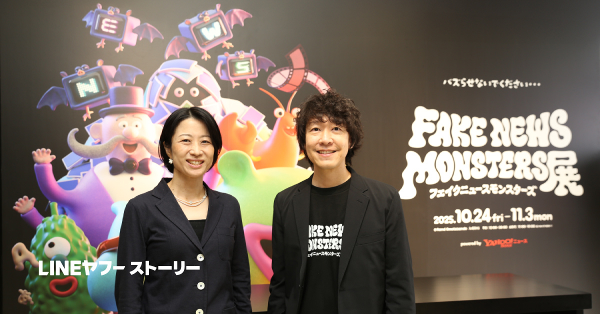 展示会「FAKE NEWS MONSTERS展」の看板前で、男女2人が並んで笑顔で立っている。背景にカラフルなモンスターのイラストが描かれている
