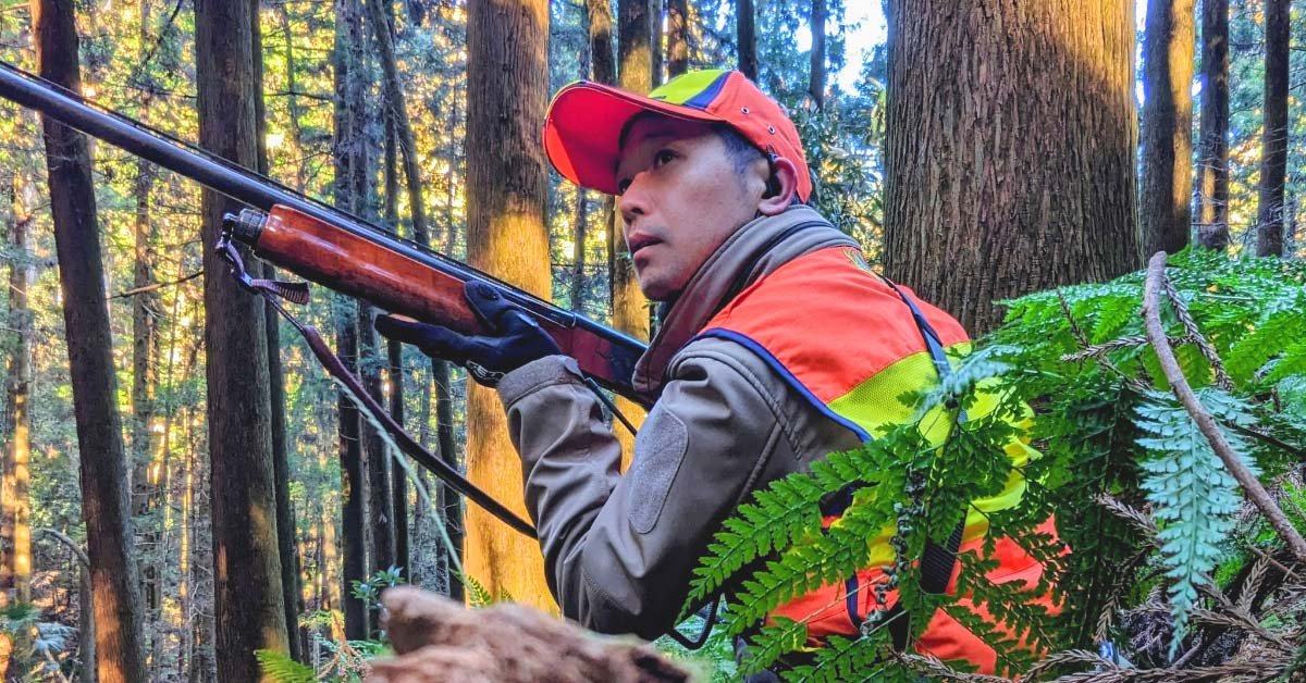 里山の木々に囲まれた場所で、猟銃を構えて周囲を確認する小山