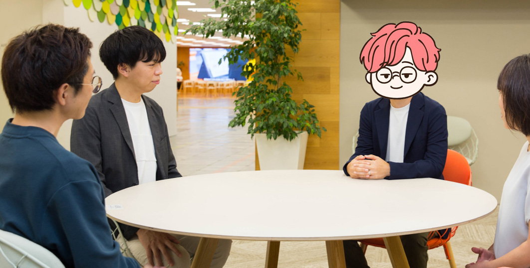 明るいオフィスの一角で、丸い白いテーブルを囲んで4人が座っている。背景には観葉植物や木目の壁、天井には緑と黄色の装飾が並んでおり、柔らかい雰囲気のオフィス空間になっている。