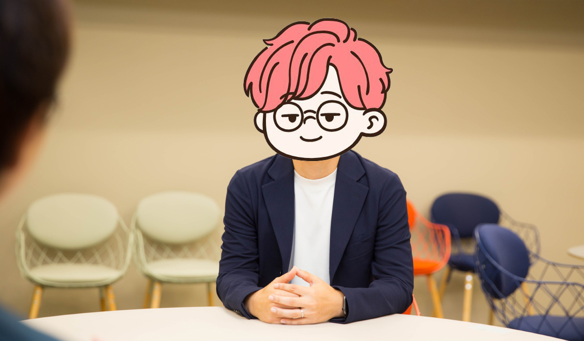 ピンク髪のキャラクター風イラストで顔を覆った男性が、オフィスの丸テーブルで話している様子。紺色のジャケットを着ている。