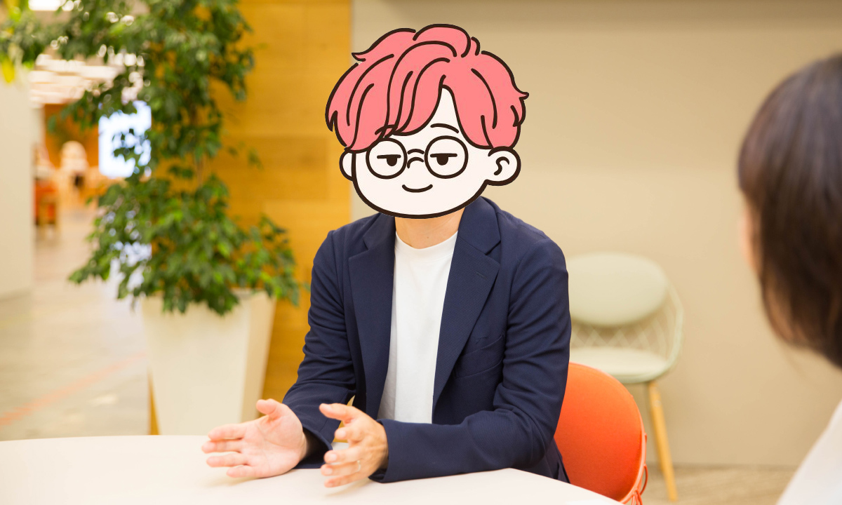 ピンク髪のキャラクター風イラストで顔を覆ったシバウラさんの写真。白いシャツに紺色のジャケットを着て、テーブルの前で手振りを交えながら話している。