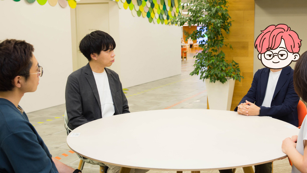 明るいオフィスの一角で、丸い白いテーブルを囲んで4人が座っている。全員が会話している様子。背景には観葉植物と木目調の壁、天井には緑や黄色の装飾が見える。