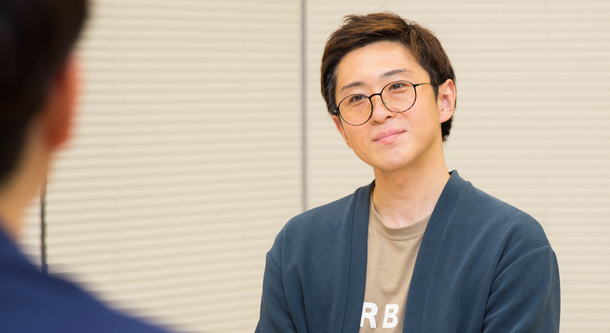 丸い眼鏡をかけた男性のバストショット。ベージュのTシャツの上に濃い青のカーディガンを羽織り、柔らかい表情で前を向いている。背景は室内で、ブラインドが閉じられた壁が見える。手前には別の人物の肩が少し写り込んでいる。