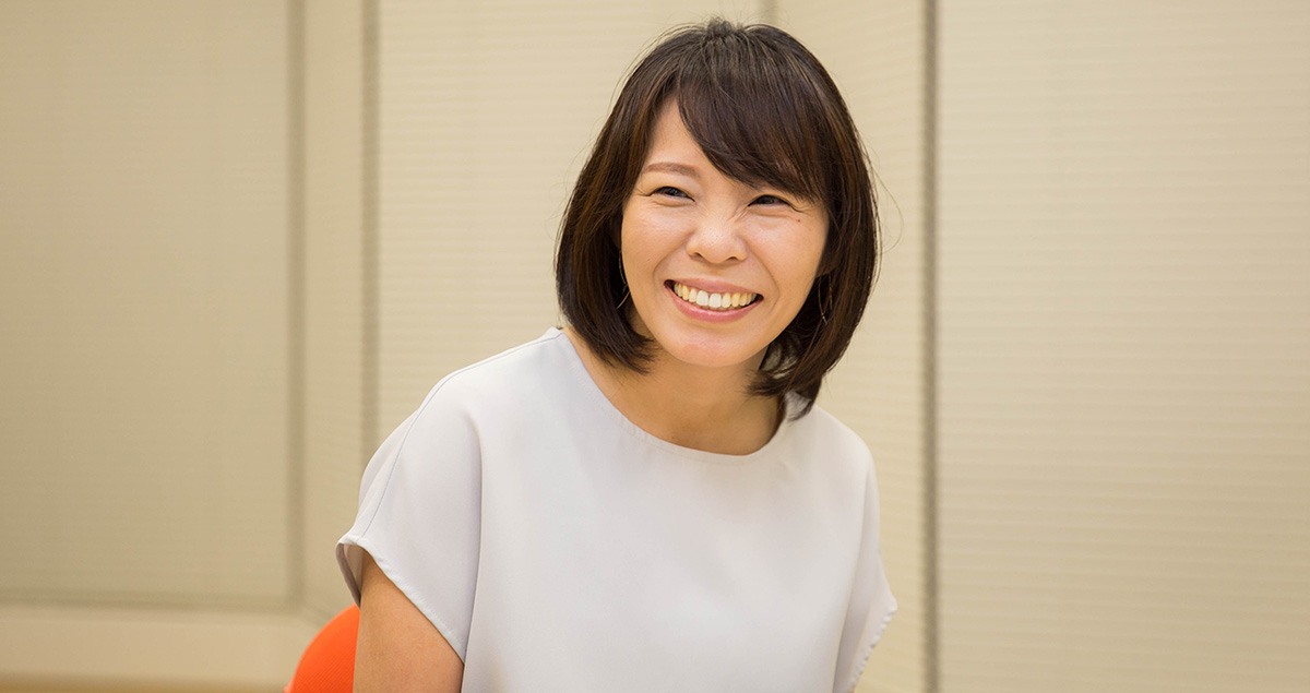 蓮池の写真。肩までの髪型で、白っぽい半袖のブラウスを着て、にこやかに笑顔を見せている。背景にはベージュのブラインドが閉じられた壁があり、柔らかい照明で全体が明るく映っている。