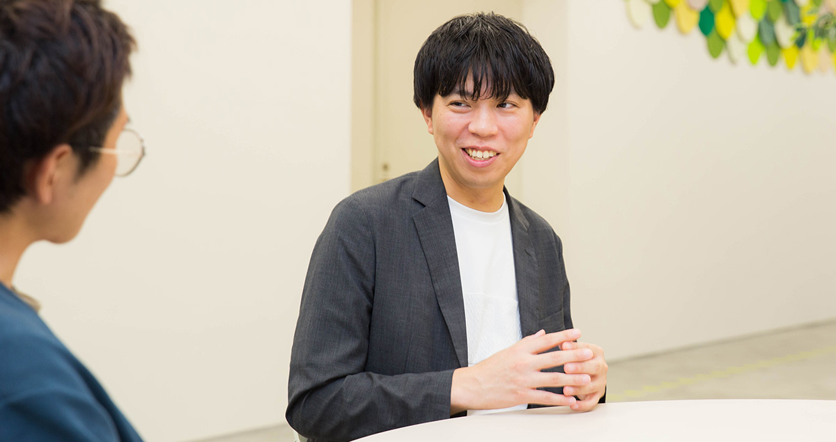 、グレーのジャケットに白いシャツを着た荻野が笑顔で会話している様子。両手を軽く組みながら向かいの人物に話しかけており、背景には白い壁と天井から吊るされた緑や黄色の装飾が見える。