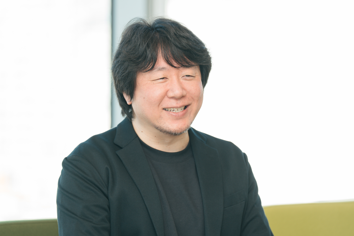 冨川のバストショット。黒のジャケットと黒のTシャツを着ており、笑顔で少し右側を向いている。