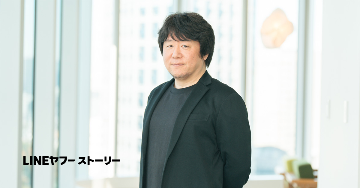 冨川のバストショット。黒のジャケットと黒のTシャツを着て静かに微笑みながらカメラを見つめている。""