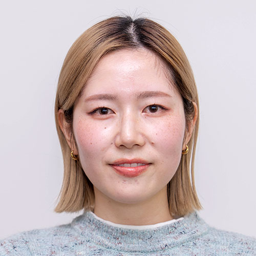 グレー背景の前で正面を向く藤目のバストショット。肩までの金髪ボブヘアで、淡いグレーのニットを着用し、小さなゴールドのピアスをつけて穏やかに微笑んでいる