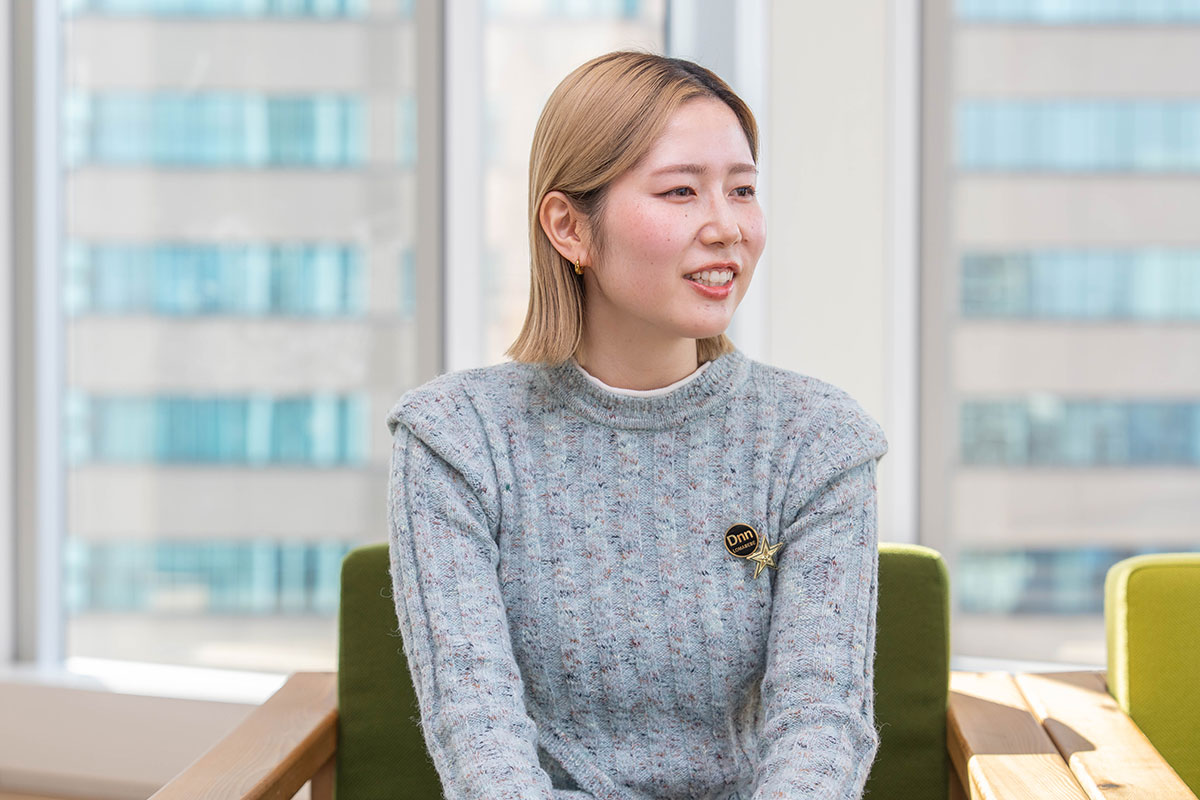 窓を背に椅子へ座る藤目の写真。淡いグレーのニットを着て横を向き、柔らかく話している様子である。背後には高層ビルが見える